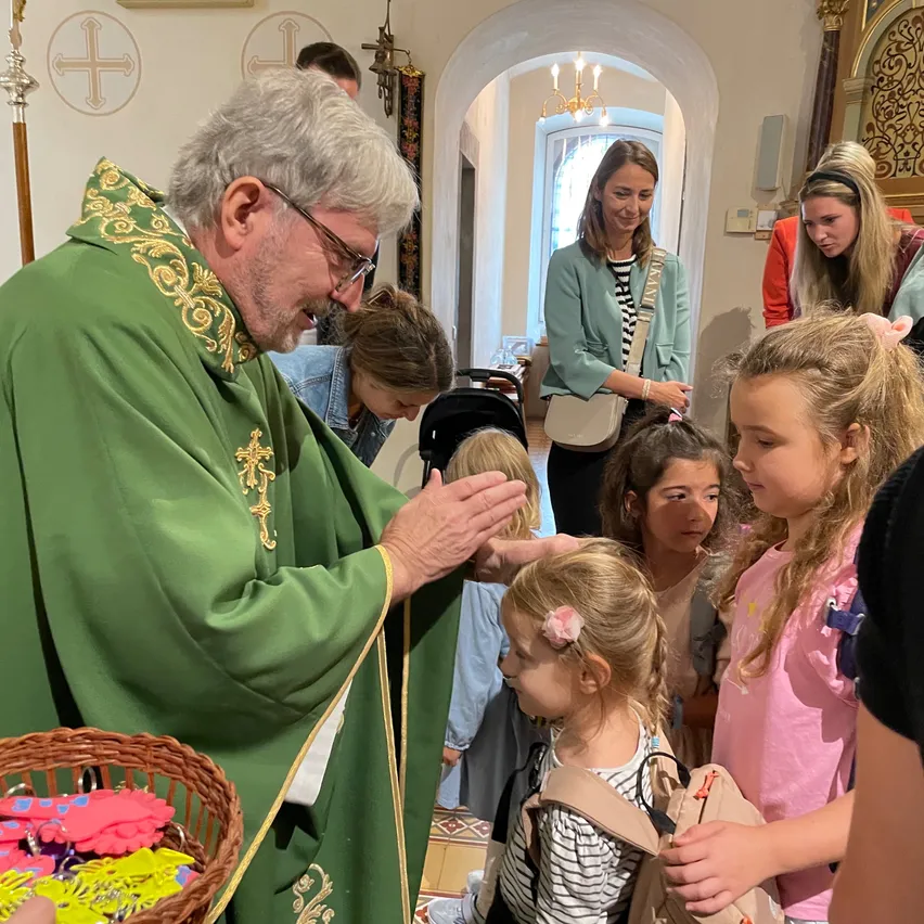 Ein Priester in einer grünen Robe segnet eine Gruppe junger Mädchen in einer Kirche, von denen eine ein Korb mit bunten Schlüsselanhängern hält.