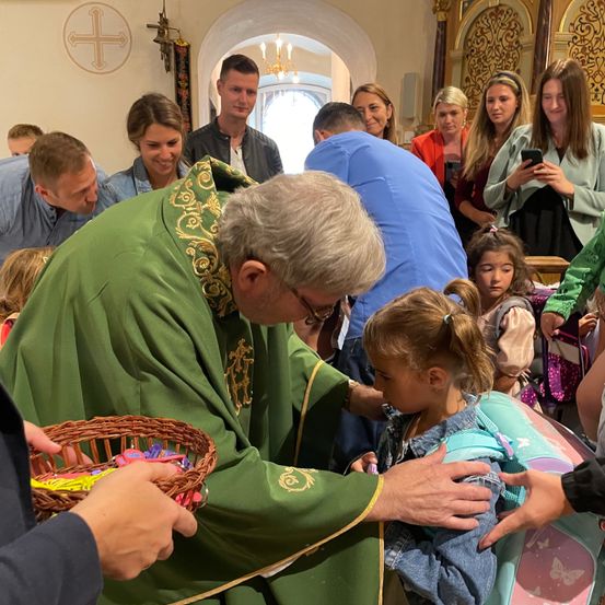 Ein Priester in einem grünen Gewand segnet ein junges Mädchen in einer Kirche. Erwachsene und Kinder versammeln sich um sie, einige halten Körbe mit Süßigkeiten. Eine Frau im Hintergrund hält den Moment mit ihrem Handy fest.