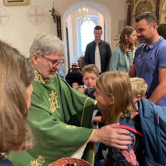 Ein Priester in einer grünen Robe segnet ein Kind in einer Kirche. Erwachsene und Kinder versammeln sich im Hintergrund, einige mit lächelnden Gesichtern.