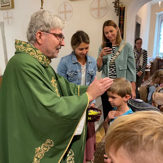 Ein Priester in einer grünen Robe segnet einen jungen Jungen, der einen Korb hält. In der Nähe macht eine Frau ein Foto mit ihrem Handy.