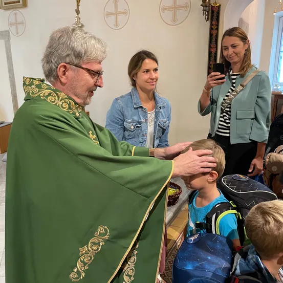 Ein Priester in einem grünen Gewand segnet einen jungen Jungen in einer Kirche. Zwei Frauen stehen hinter ihnen, eine macht ein Foto mit ihrem Handy.