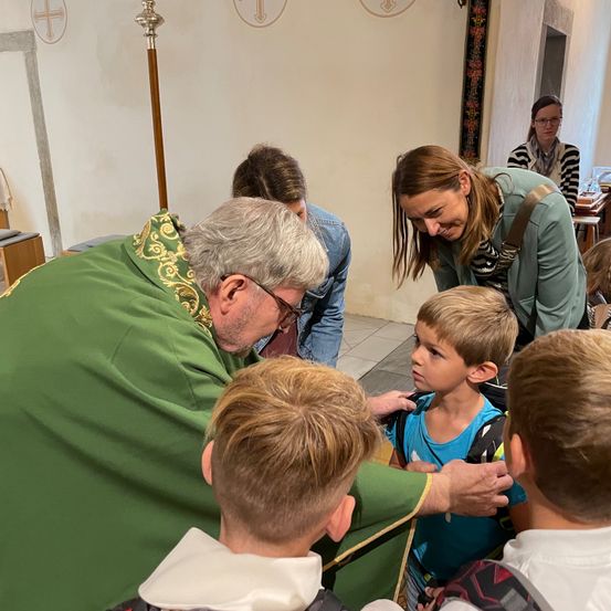 Ein Priester in grüner Amtstracht segnet junge Kinder, die Rucksäcke tragen. Eine Frau beobachtet, während andere im Hintergrund sitzen.