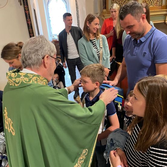 Ein Priester in einer grünen Robe segnet einen Jungen in einer Kirche, umgeben von Familien.