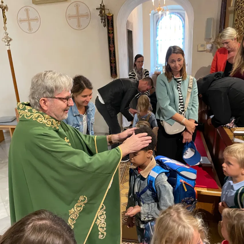 Ein Priester segnet den Kopf eines Kindes in einer Kirche. Andere Leute stehen und beobachten. Ein Mädchen hält einen Rucksack.
