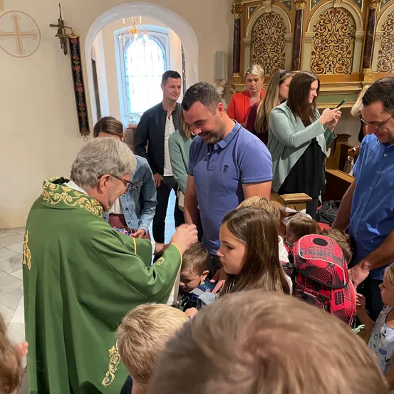 In einer Kirche segnet ein Priester in grünen Gewändern eine Gruppe von Kindern, während Erwachsene, einige mit Brille, zusehen. Die Szene spielt sich in der Nähe eines Bogens mit einem Kreuz ab.
