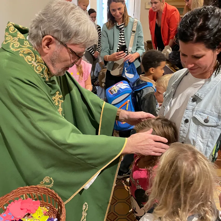 Ein Priester segnet Kinder in einer Kirche, umgeben von Familien. Kinder halten Rucksäcke, und Erwachsene interagieren mit ihnen.