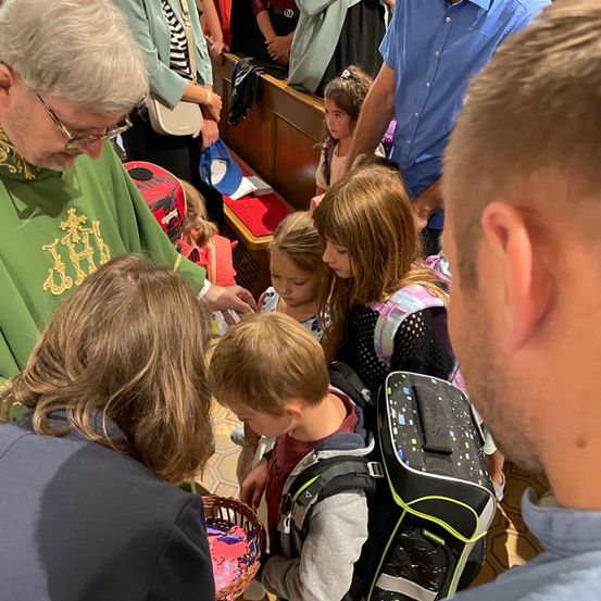 Ein Priester in grünen Gewändern verteilt Gegenstände an Kinder in einer Kirchenumgebung, mit Erwachsenen und Kindern, die sich versammelt haben, einige mit Rucksäcken.