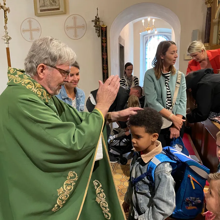 Ein Priester in grüner Gewandung segnet ein Kind mit einem Rucksack während einer Kirchenzeremonie, mit Erwachsenen und Kindern in der Umgebung.