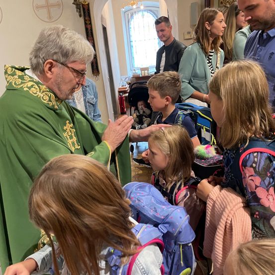 Ein Priester in grüner Robe segnet Kinder in einer Kirche. In der Nähe warten Eltern und Kinder mit Rucksäcken.