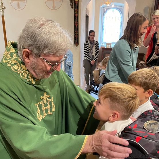 Ein Priester in einer grünen Robe segnet zwei Jungen. Eine Frau und ein Mädchen beobachten im Hintergrund.