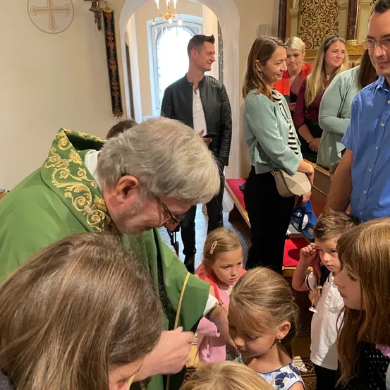 Ein Priester in grünen Gewändern spendet Kindern in einer Kirche einen Sakrament. Mehrere Erwachsene stehen in der Nähe und unterhalten sich. Ein Kreuz ist an der Wand zu sehen.