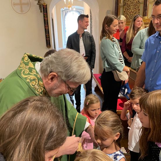 Ein Priester in grünen Gewändern spendet Kindern in einer Kirche einen Sakrament. Mehrere Erwachsene stehen in der Nähe und unterhalten sich. Ein Kreuz ist an der Wand zu sehen.