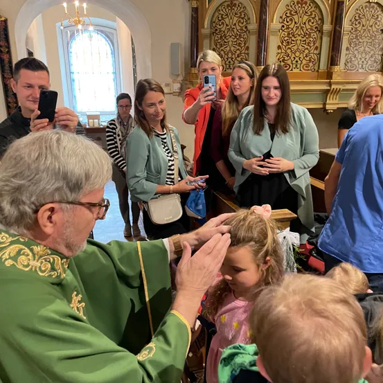 Ein Priester in grünen Gewändern segnet ein junges Mädchen in einer Kirche. Mehrere Menschen sind anwesend, darunter ein Mann, der ein Foto macht, und andere, die beobachten.
