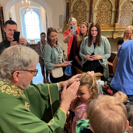 Ein Priester in grünen Gewändern segnet ein junges Mädchen in einer Kirche. Mehrere Menschen sind anwesend, darunter ein Mann, der ein Foto macht, und andere, die beobachten.