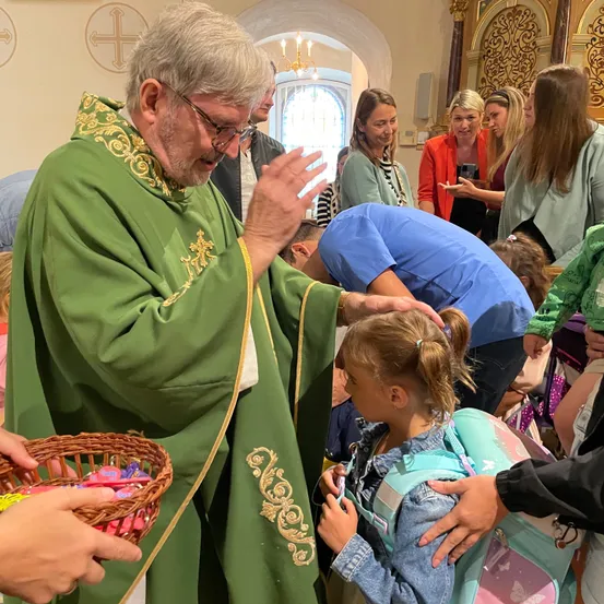 Ein Priester in grünen Gewändern segnet ein junges Mädchen mit Rucksack in einer Kirche, während Erwachsene und Kinder um sie herum versammelt sind.