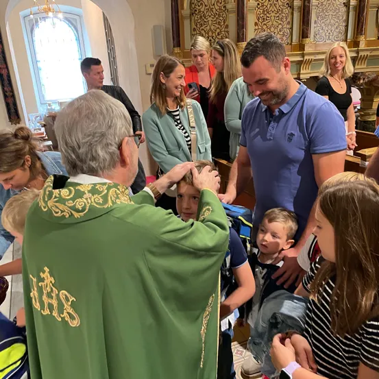 Ein Priester in einem grünen Gewand segnet ein junges Kind, während Erwachsene und Kinder zusehen. Der Schauplatz ist eine Kirche mit hohen Decken und einem Buntglasfenster.