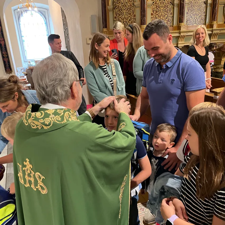 Ein Priester in einem grünen Gewand segnet ein junges Kind, während Erwachsene und Kinder zusehen. Der Schauplatz ist eine Kirche mit hohen Decken und einem Buntglasfenster.