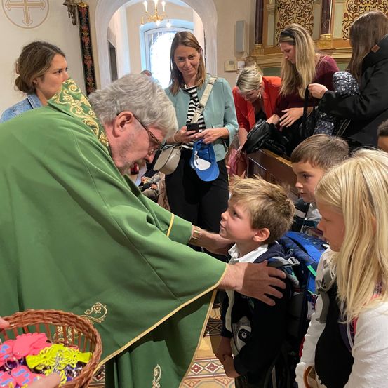 Ein Priester in einem grünen Gewand segnet einen jungen Jungen mit einem Rucksack, umgeben von Erwachsenen und Kindern in einer Kirchenumgebung.