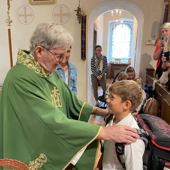 Ein Priester tröstet einen jungen Jungen, der einen Rucksack trägt, in einer Kirche. Hinter ihnen stehen Leute.
