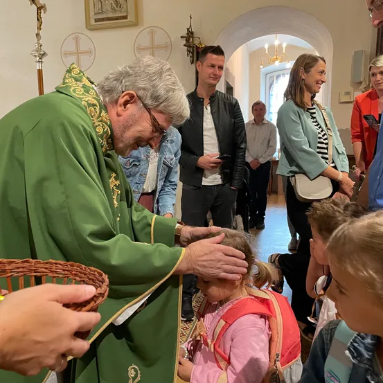 Ein Priester in grünen Gewändern segnet den Kopf eines jungen Mädchens in einer Kirche. Hinter ihnen steht eine Gruppe von Menschen und beobachtet. Ein Korb wird von einer Person auf der linken Seite gehalten.