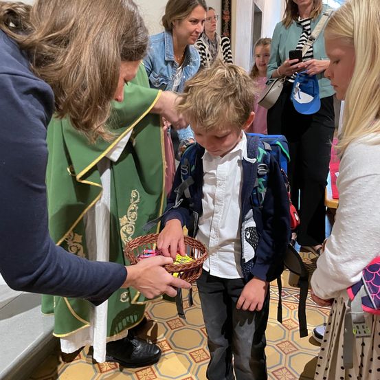 Eine Frau in einem grünen Gewand überreicht einem Jungen mit ernstem Gesicht einen Korb. Der Junge trägt einen Rucksack und hält einen Korb mit Süßigkeiten.