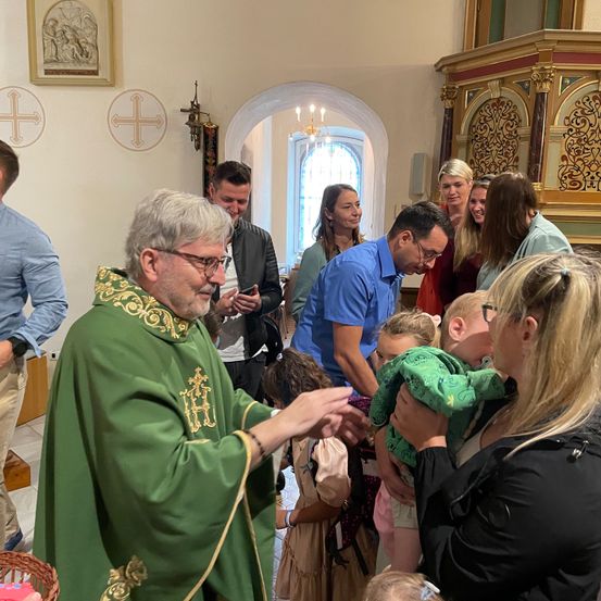 Ein Priester in grünen Gewändern interagiert mit einer Familie und ihren Kindern in einer Kirche. Der Priester hält ein Baby in seinen Armen.