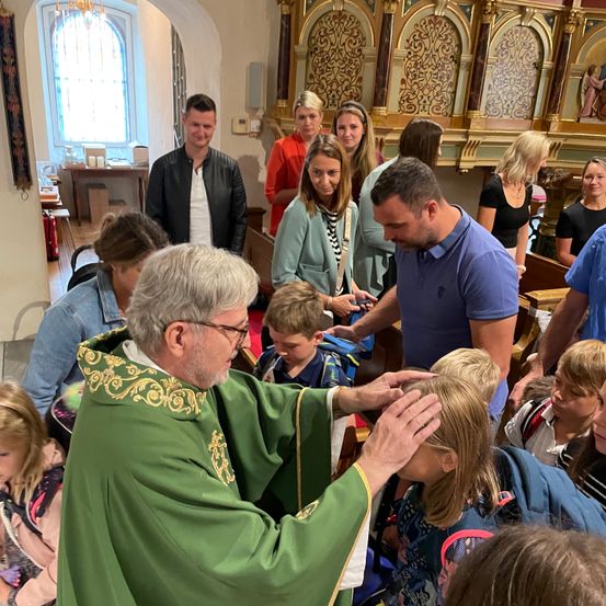 Ein Priester in einer grünen Robe segnet ein Kind in einer Kirche, während mehrere Menschen stehend zusehen.