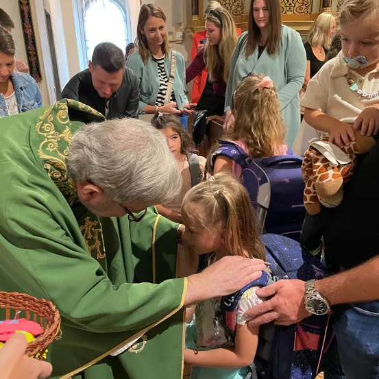 Ein Priester in grünen Gewändern segnet ein junges Mädchen mit einem Rucksack und einem Kuscheltier in einer Kirche.