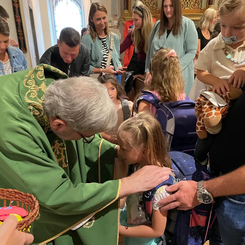Ein Priester in grünen Gewändern segnet ein junges Mädchen mit einem Rucksack und einem Kuscheltier in einer Kirche.