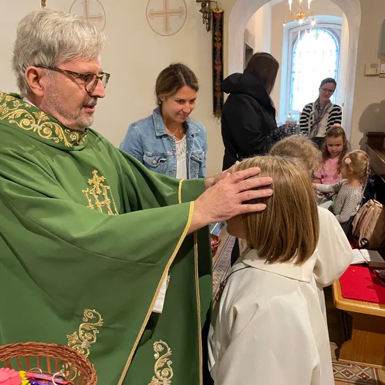 Ein Priester in einem grünen Gewand mit goldenen Verzierungen segnet ein junges Mädchen in einem weißen Gewand. Hinter ihnen beobachtet eine Gruppe von Menschen, darunter eine Frau in einer Jeansjacke und ein Mädchen in einem rosa Shirt.