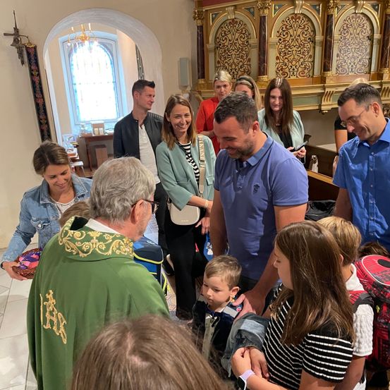 Eine Gruppe von Menschen, darunter Erwachsene und Kinder, versammelt sich in einer Kirche. Sie alle lächeln und schauen zu einem Priester in einem grünen Gewand. Der Priester hält einen Korb und scheint mit den Menschen zu sprechen.