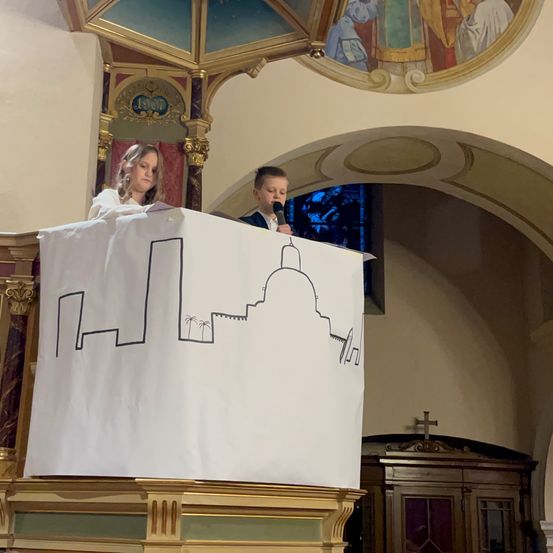 Zwei Kinder stehen an einem Podium in einer Kirche, halten ein Mikrofon und ein Banner mit einer gezeichneten Stadtsilhouette. Hinter ihnen ist ein gewölbter Durchgang und ein kunstvoll verzierter hölzerner Schrank mit einem Kreuz zu sehen.