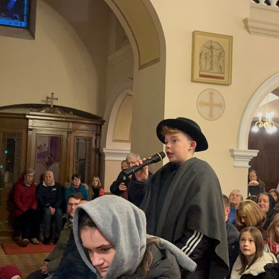 Ein Junge in Schwarz singt in ein Mikrofon in einer Kirche. Er steht vor einem sitzenden Publikum.