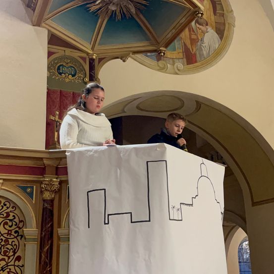 Eine Frau und ein Junge stehen hinter einem Banner mit einer Zeichnung, möglicherweise in einer Kirche, mit einem Bogen und einem Kreuz im Hintergrund.