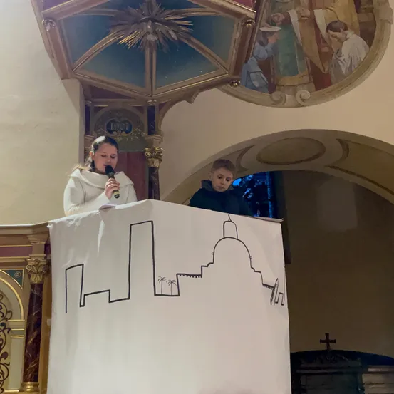 Ein Mädchen in einer weißen Robe spricht in ein Mikrofon an einem Rednerpult, mit einem Jungen dahinter. Ein Banner mit einer Kirchen-Design ist vor ihnen. Ein Wandbild mit religiösen Figuren ist darüber.