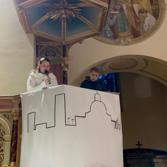 Ein Mädchen in einer weißen Robe spricht in ein Mikrofon an einem Rednerpult, mit einem Jungen dahinter. Ein Banner mit einer Kirchen-Design ist vor ihnen. Ein Wandbild mit religiösen Figuren ist darüber.