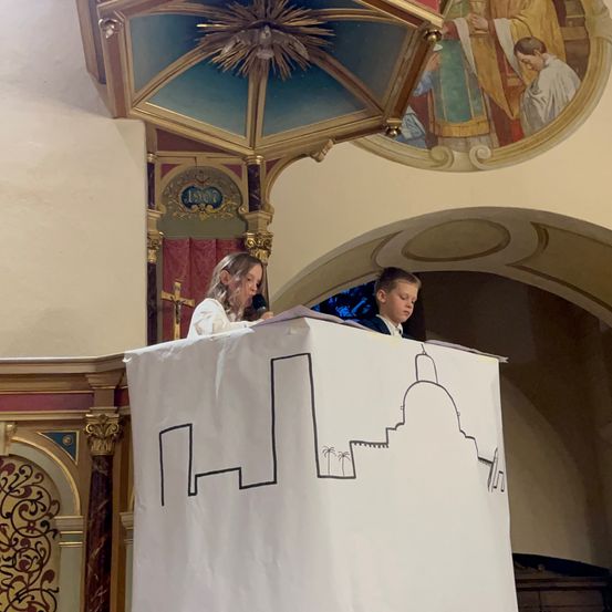 Zwei Kinder stehen hinter einem Podium mit einer Zeichnung auf einem weißen Hintergrund. Das Mädchen sieht nach unten und der Junge schaut nach vorne. Dahinter zeigt eine Wandmalerei eine religiöse Szene.