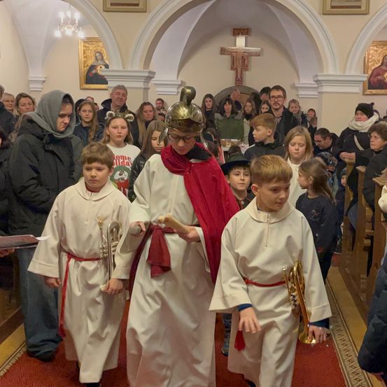 Ein Kirchenumzug mit jungen Kindern in weißen Gewändern und roten Schärpen, eines trägt eine Trompete, und eine Person in einer römischen Soldatenkostümierung führt an.