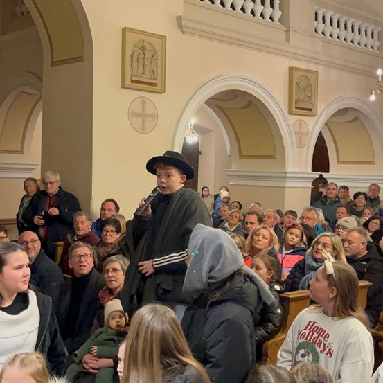 Eine Gruppe von Menschen ist in einer Kirche versammelt. Ein junger Mann in einem Hut spricht in ein Mikrofon. Das Publikum hört aufmerksam zu, und einige machen Fotos.