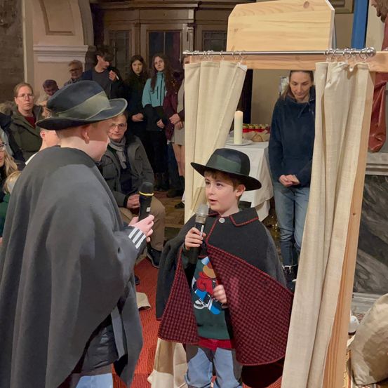 Ein Junge in einem Umhang hält ein Mikrofon und spricht, während ein Mann in einem Hut zuhört. Hinter ihnen beobachtet eine Gruppe von Menschen, und eine Frau steht in der Nähe eines Tisches mit einer Kerze.
