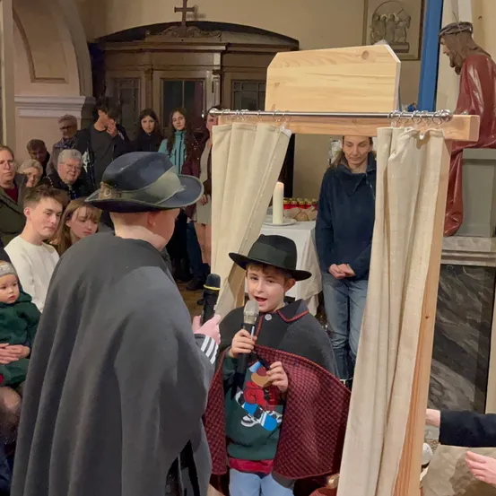 Ein Junge spricht in ein Mikrofon in einer Kirche, während die Menschen im Hintergrund zusehen. In der Nähe befindet sich eine Holzkonstruktion mit Vorhängen und einer Kerze.