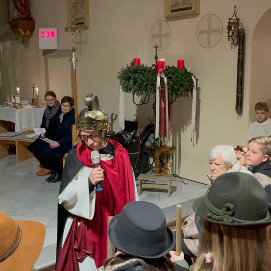 Eine Person in mittelalterlicher Kleidung spricht in ein Mikrofon vor einem Publikum. Dahinter sitzen zwei Frauen auf einer Bank mit Kerzen und einem Weihnachtskranz.