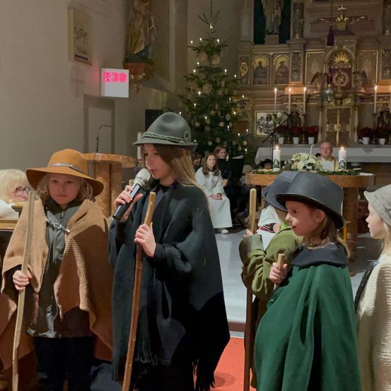 Mehrere Kinder in Kostümen stehen in einer Kirche, eines hält ein Mikrofon und spricht. Die Kinder halten Stöcke und tragen Hüte. Hinter ihnen steht ein Altar mit Kerzen und einem Weihnachtsbaum.