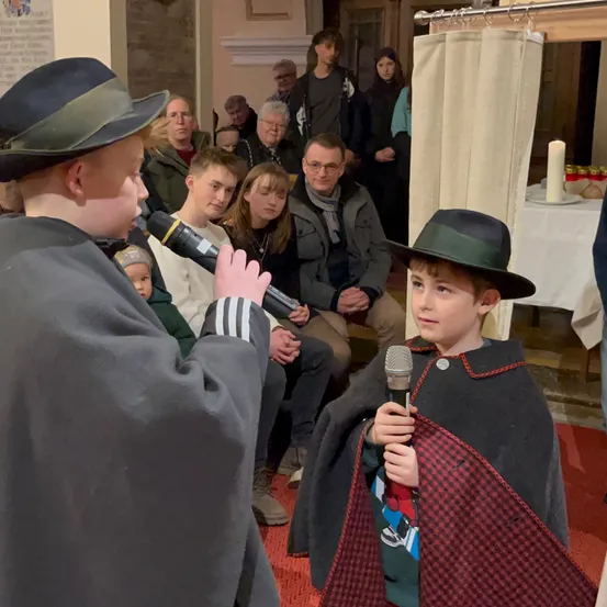 Zwei Jungen, einer in einem schwarzen Umhang und der andere in einem grauen Mantel, halten Mikrofone und sprechen in einem Raum mit einem sitzenden Publikum. Dahinter sind eine Kerze und ein Tisch mit Tellern und Flaschen zu sehen.