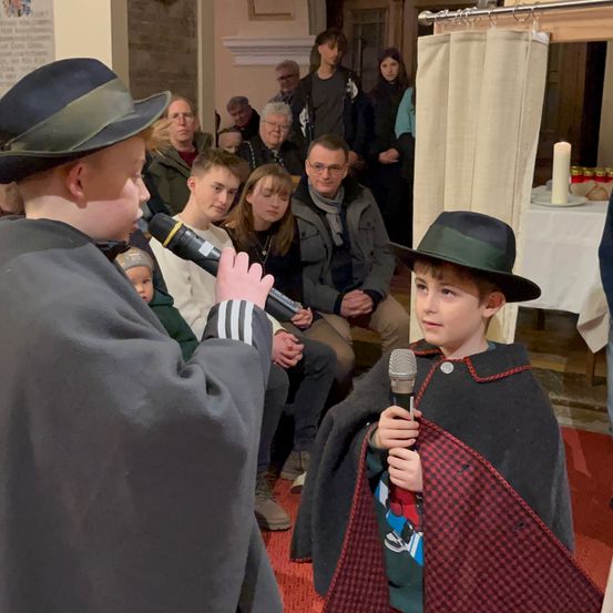 Zwei Jungen, einer in einem schwarzen Umhang und der andere in einem grauen Mantel, halten Mikrofone und sprechen in einem Raum mit einem sitzenden Publikum. Dahinter sind eine Kerze und ein Tisch mit Tellern und Flaschen zu sehen.