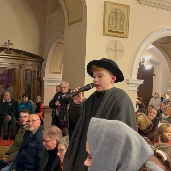 Ein junger Junge in einem schwarzen Hut und Umhang singt in ein Mikrofon in einer Kirche, mit einem Publikum von sitzenden und stehenden Menschen hinter ihm.