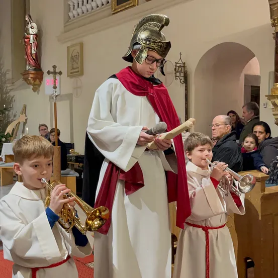 In einer Kirche liest ein junger Mann mit Helm aus einem Buch. Zwei junge Jungen, ebenfalls in Weiß, spielen Trompeten. Eine Statue und ein Kreuz sind im Hintergrund.