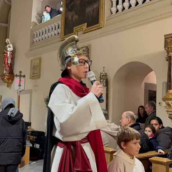 Ein Junge in einem Kostüm mit Helm und Brille spricht in ein Mikrofon in einer Kirche, während ein kleiner Junge zuhört. Mehrere Menschen sehen von den Kirchenbänken zu.