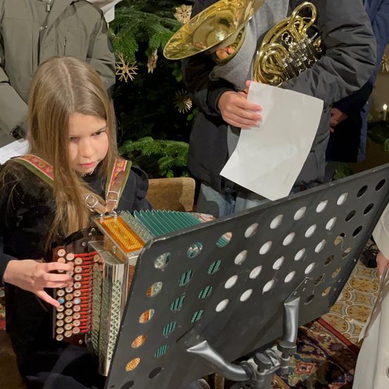 Ein junges Mädchen spielt Akkordeon bei einem Feiertagsereignis, begleitet von einer Person mit Notenpapier und einem Blechblasinstrument.