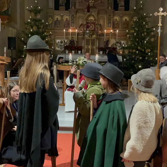 In einer Kirche stehen mehrere Kinder und Erwachsene in mittelalterlicher Kleidung. Sie stehen in der Nähe eines Altars mit brennenden Kerzen und Blumen. Die Umgebung umfasst Weihnachtsbäume und ein Kreuz auf einem Ständer.